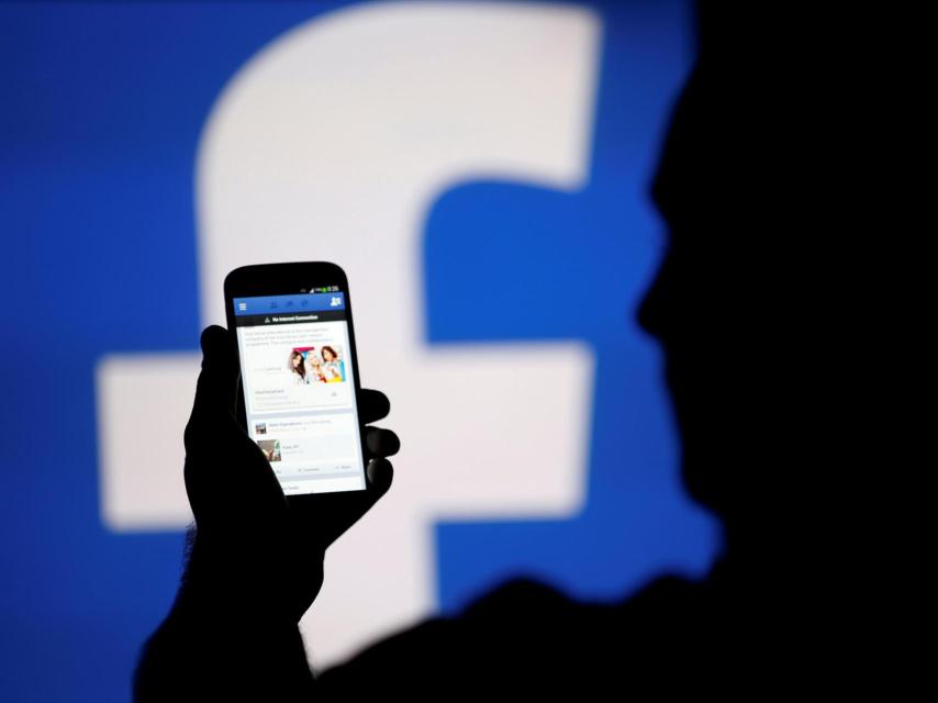 Un hombre consulta su móvil frente al logo de Facebook