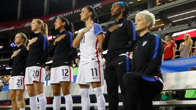 Megan Rapinoe escucha el himno nacional con una rodilla en el suelo.