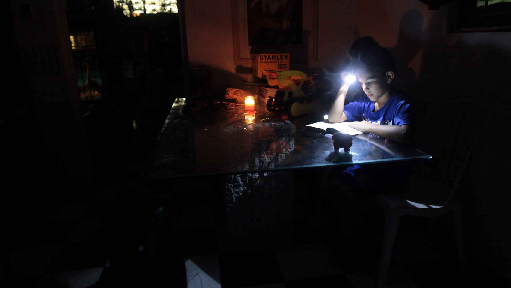 Una niña lee un libro alumbrándose con una linterna en la oscuridad de su casa, que permanece sin electricidad