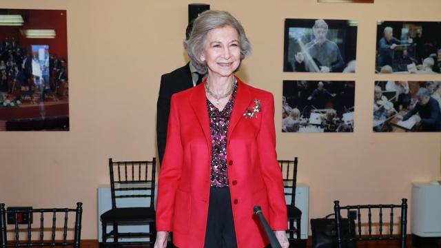 La reina Sofía luciendo un pantalón de pinzas negro y una blusa estampada en tonos morados.