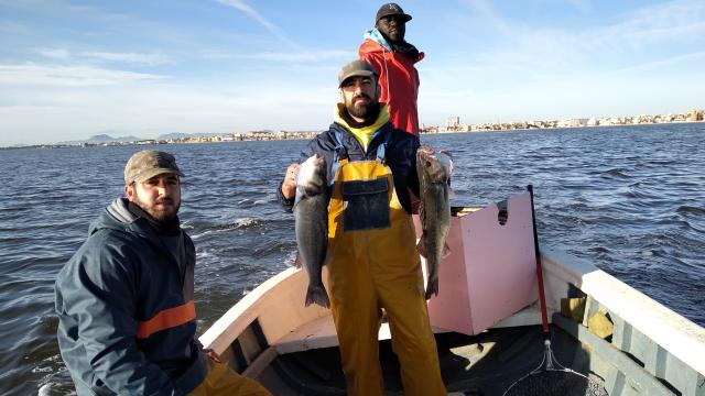 José, Myacine y el patrón de la 'Begoñita', Manolo, mostrando dos lubinas capturadas en el Mar Menor cuyas aguas se ven turbias al fondo.