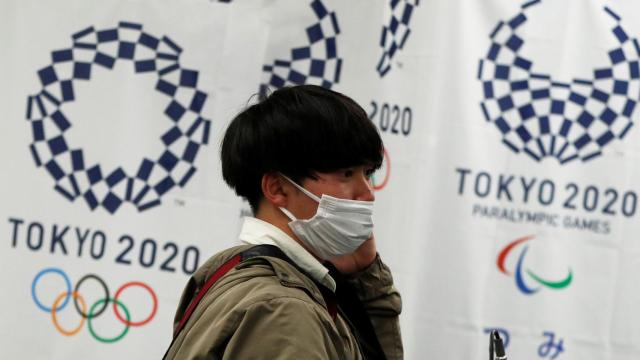 Un joven japonés con mascarilla, delante del logo de Tokio 2020