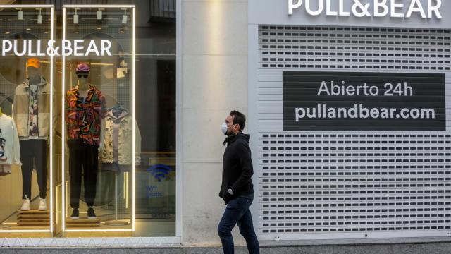 Una persona con mascarilla camina frente a una tienda cerrada y vacía.