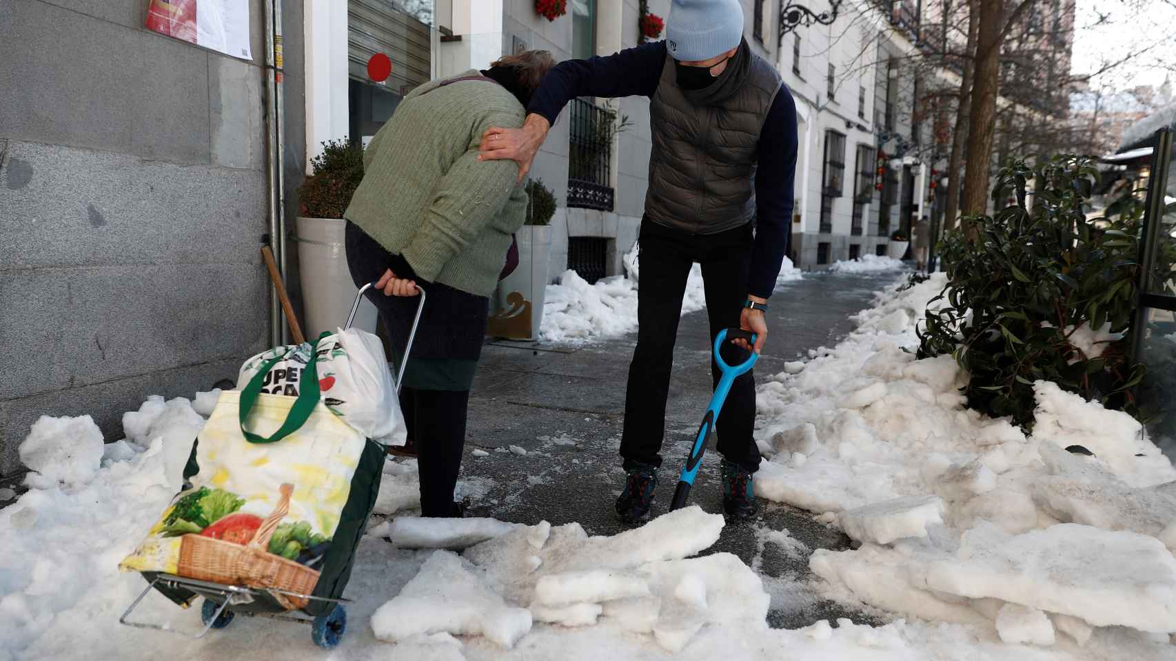 Una mujer camina con un carro de la compra mientras un hombre retira nieve de la acera este lunes en Madrid.