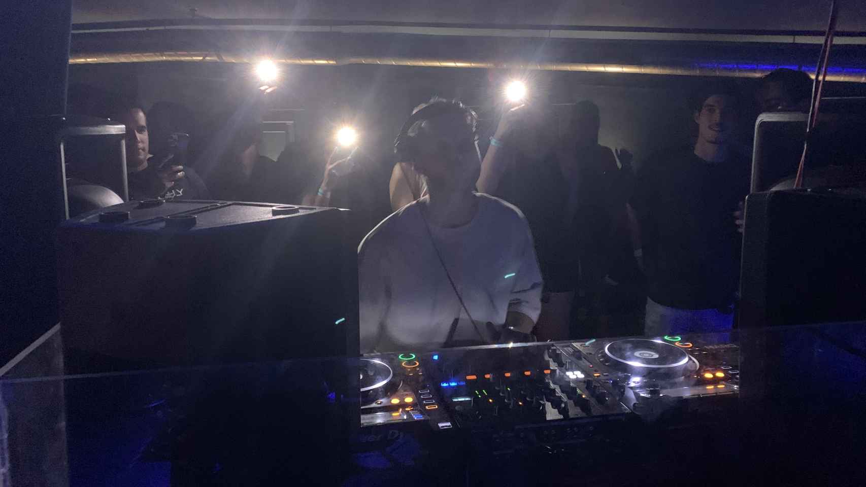 Un DJ pincha música electrónica en una discoteca. Tras él, un reservado abarrotado.