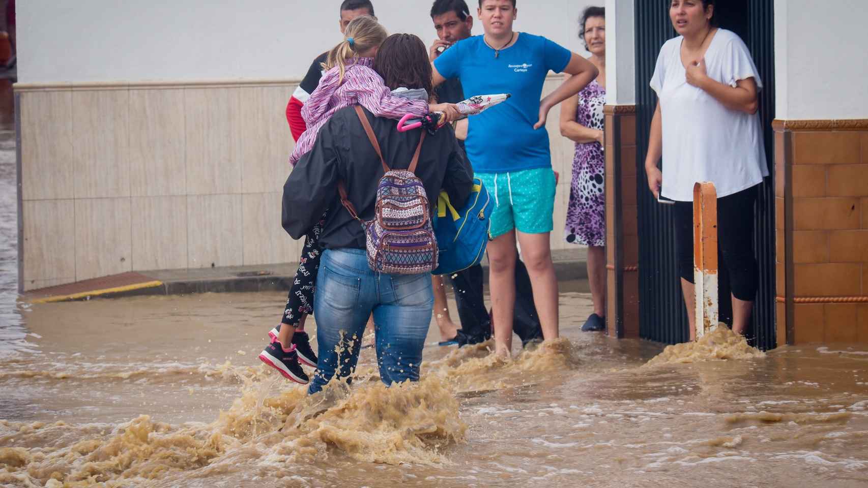 Vecinos de Lepe (Huelva) observan a una mujer con una niña en brazos cruzando una calle inundada tras las fuertes lluvias.