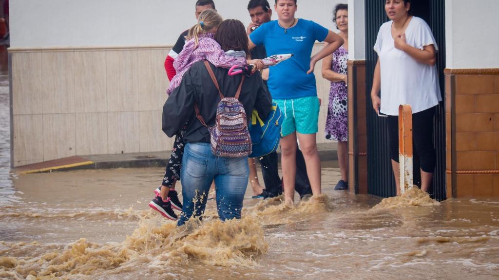 Vecinos de Lepe (Huelva) observan a una mujer con una niña en brazos cruzando una calle inundada tras las fuertes lluvias.