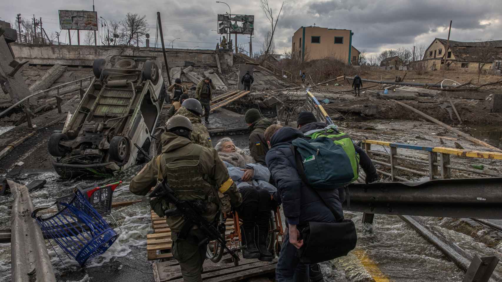 Miembros del ejército ucraniano junto con otras personas llevan a una anciana en silla de ruedas para cruzar el puente destruido mientras la gente huye de la ciudad de Irpin, en la región de Kyiv (Kiev).