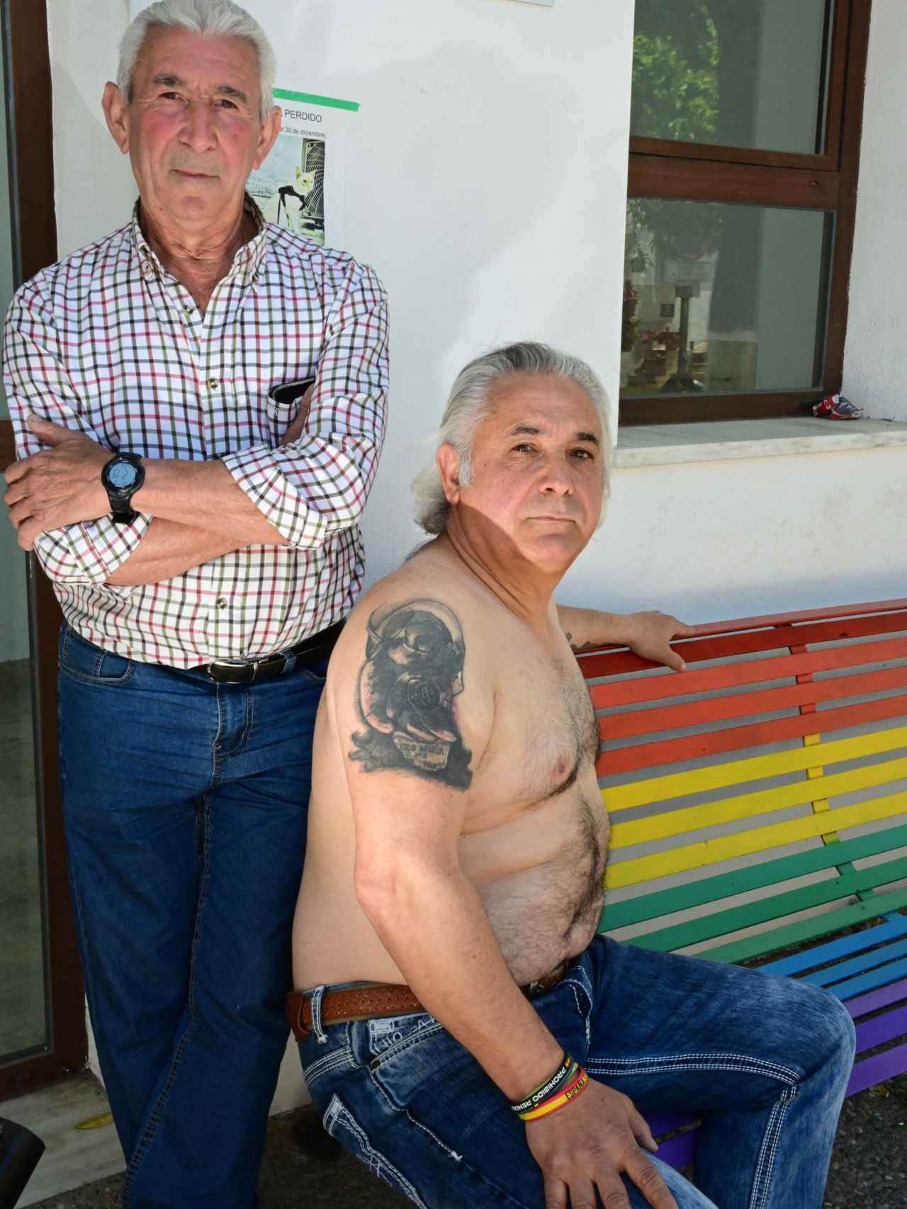 José María y Daniel posan en el centro del pueblo,  en el banco con los colores del arcoíris.