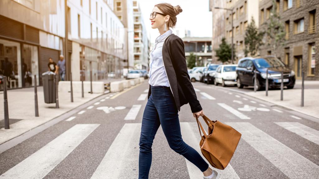 Una mujer camina hacia su oficina vestida con una americana, camisa blanca y vaqueros.