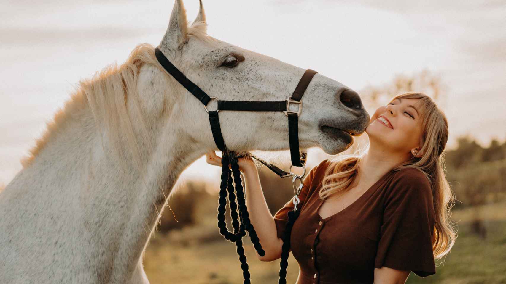 Una mujer rubia mimándose con un caballo blanco.