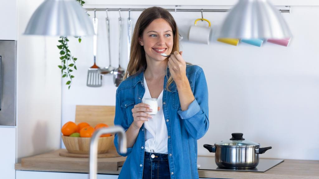 Una mujer joven comiendo yogur mientras está de pie en la cocina de casa