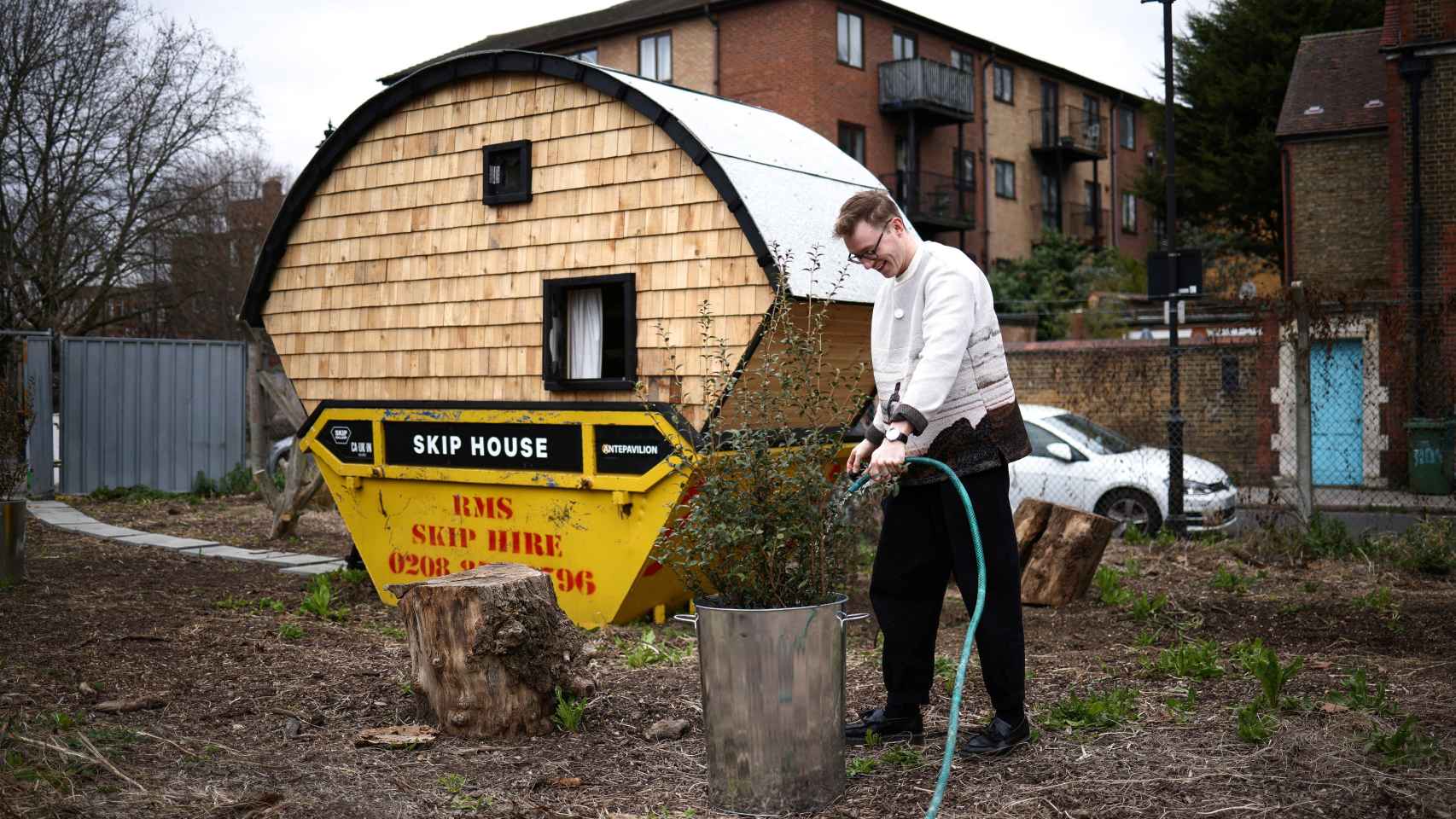 El artista Harrison Marshall riega una planta fuera del contenedor que ha convertido en vivienda, donde pretende vivir durante un año,.
