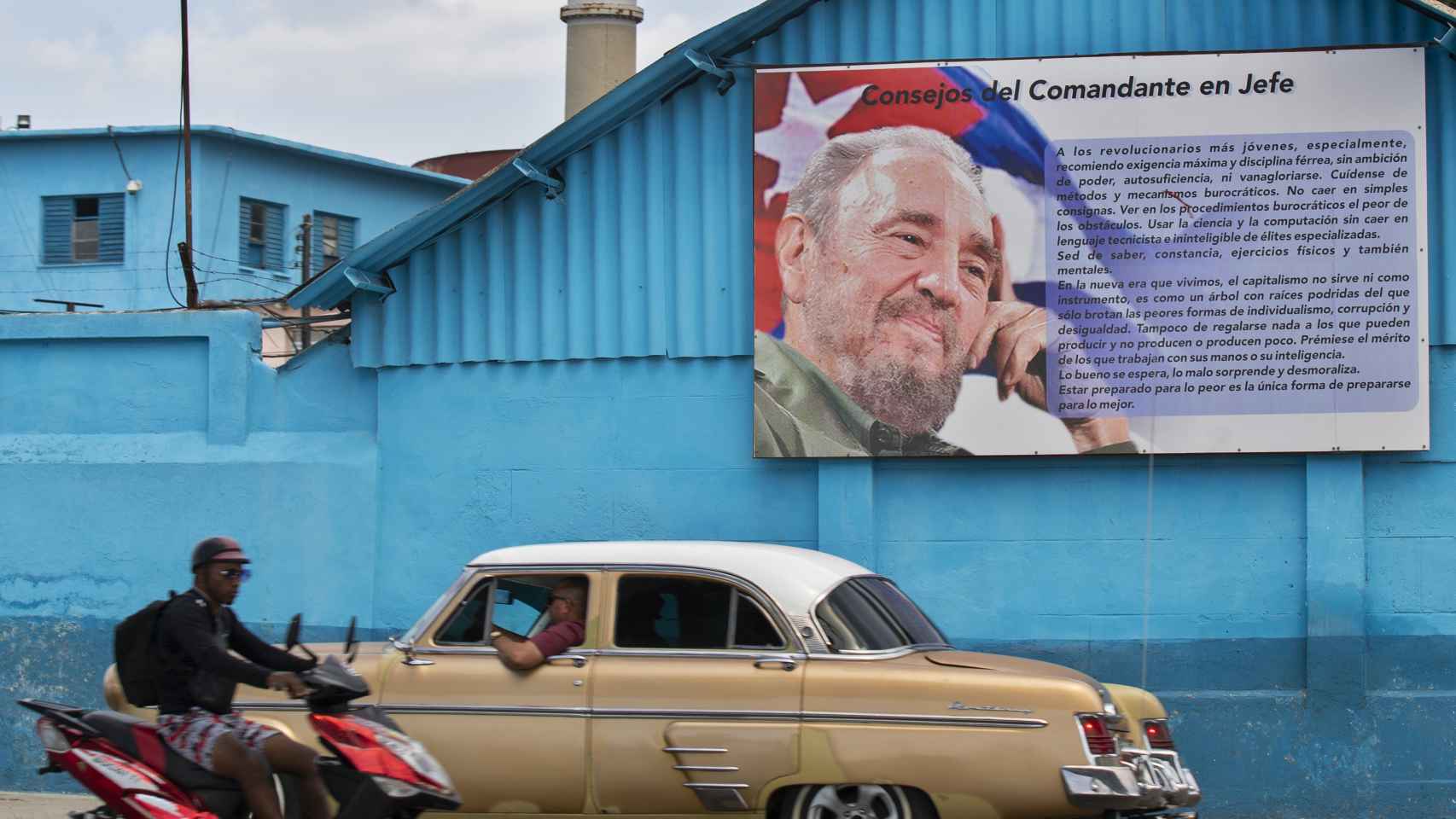 Un auto clásico y una motocicleta pasan frente a una valla con un retrato de Fidel Castro, este domingo.