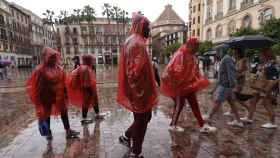 Un grupo de personas se tapan de la lluvia con chubasqueros y paraguas mientras caminan por una calle de Málaga.