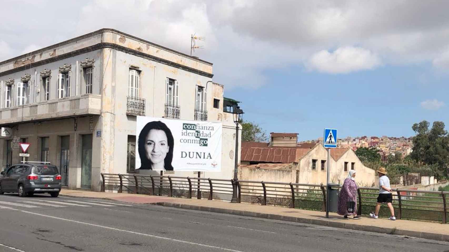 Personas pasean por una calle de Melilla junto a un cartel electoral de Coalición por Melilla.