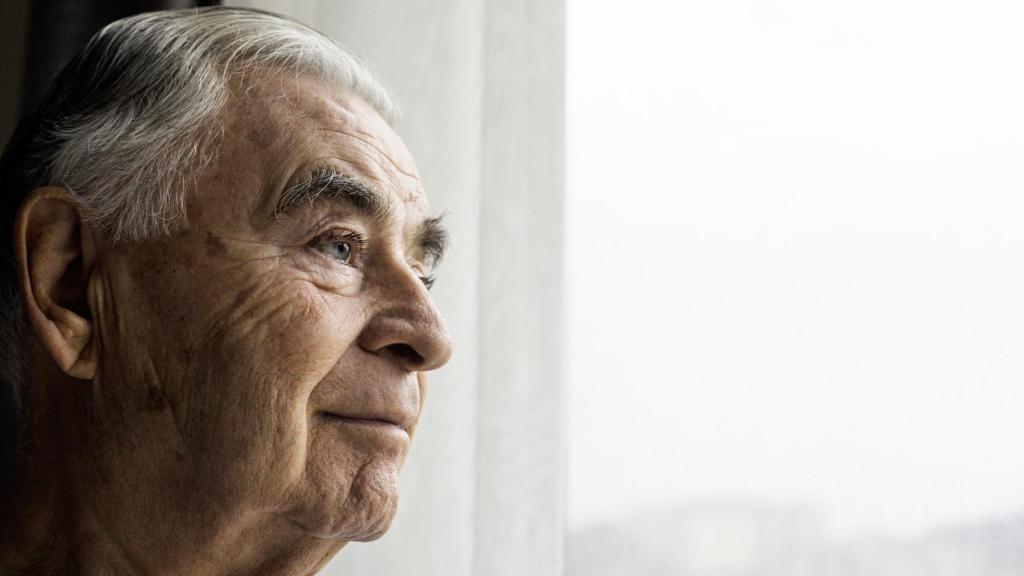 Un hombre de edad avanzada mira por la ventana de su hogar.