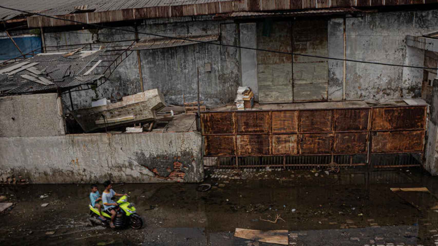 Los niños en moto pasan por un antiguo almacén detrás del malecón en la zona de Muara Baru, en el norte de Yakarta.