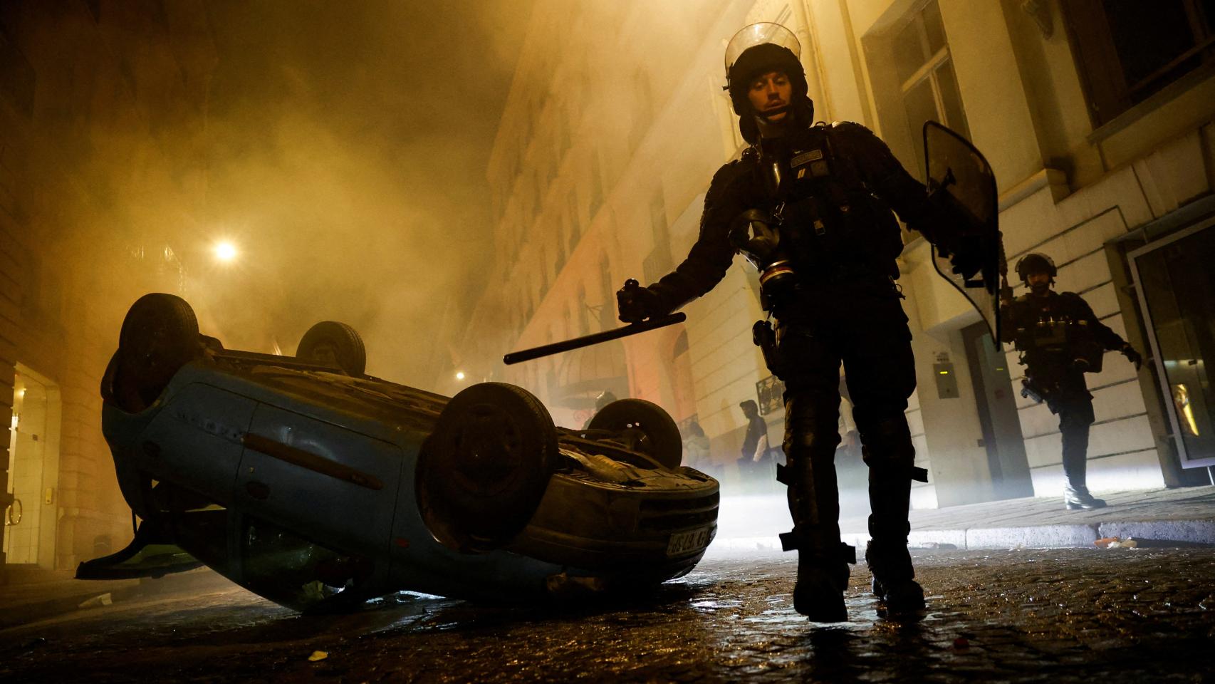 Agentes antidisturbios franceses caminan junto a un vehículo volcado durante el quinto día de protestas.
