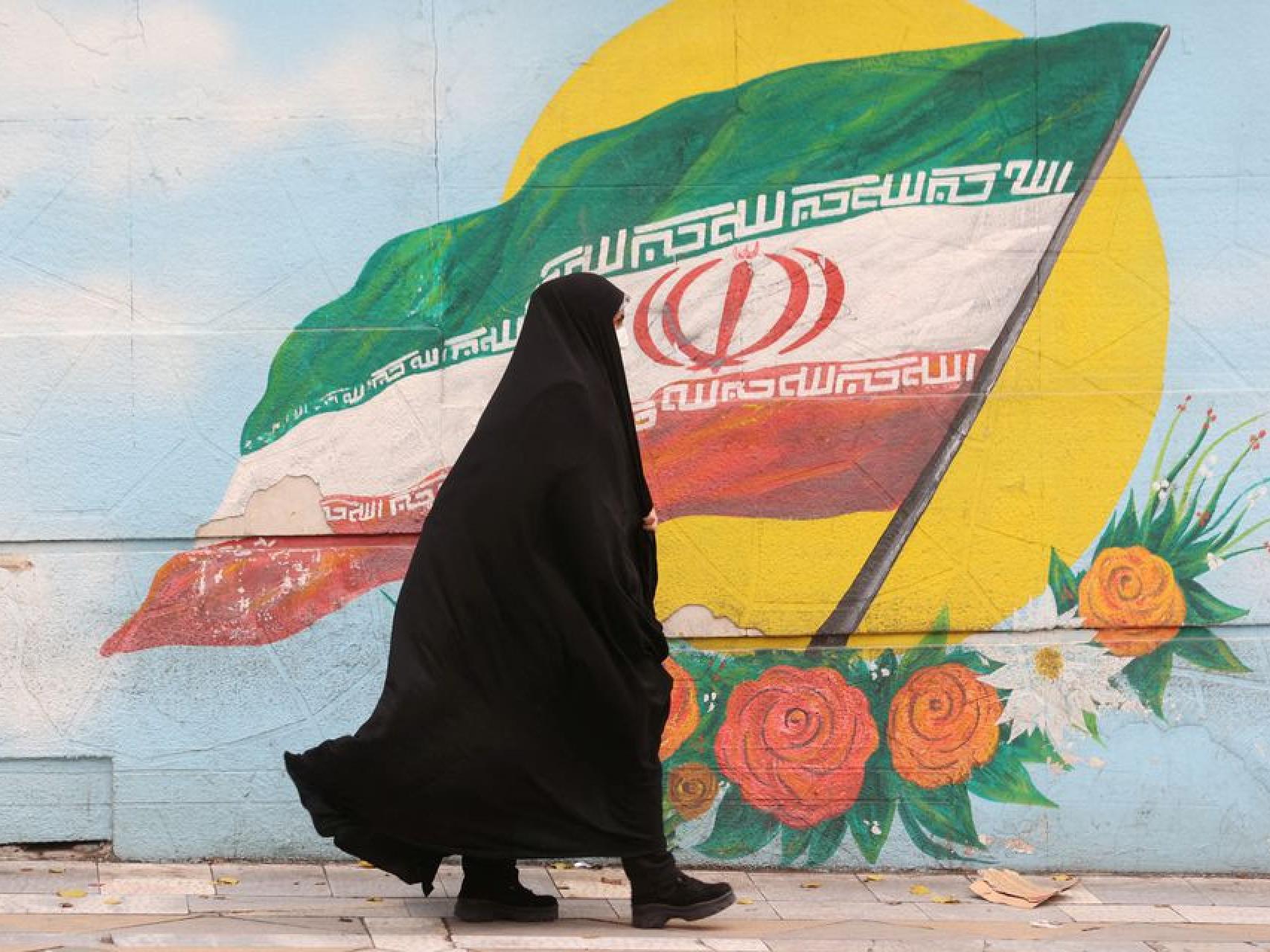 Una mujer pasea por las calles de Teherán con un mural de la bandera islámica de fondo.