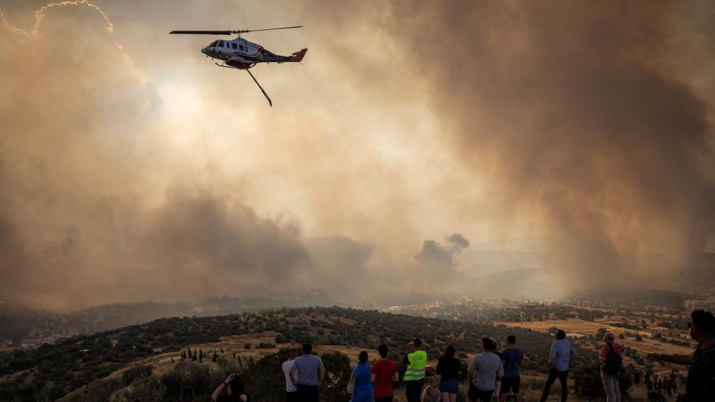 Un grupo de personas observan al helicóptero de los bomberos intentando sofocar las llamas en Mandra, Grecia.