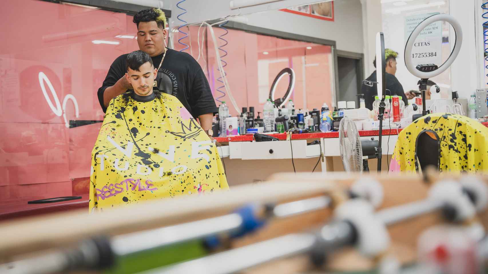 Anthony Márquez cortando el pelo a un cliente con el futbolín de la barbería asomando.