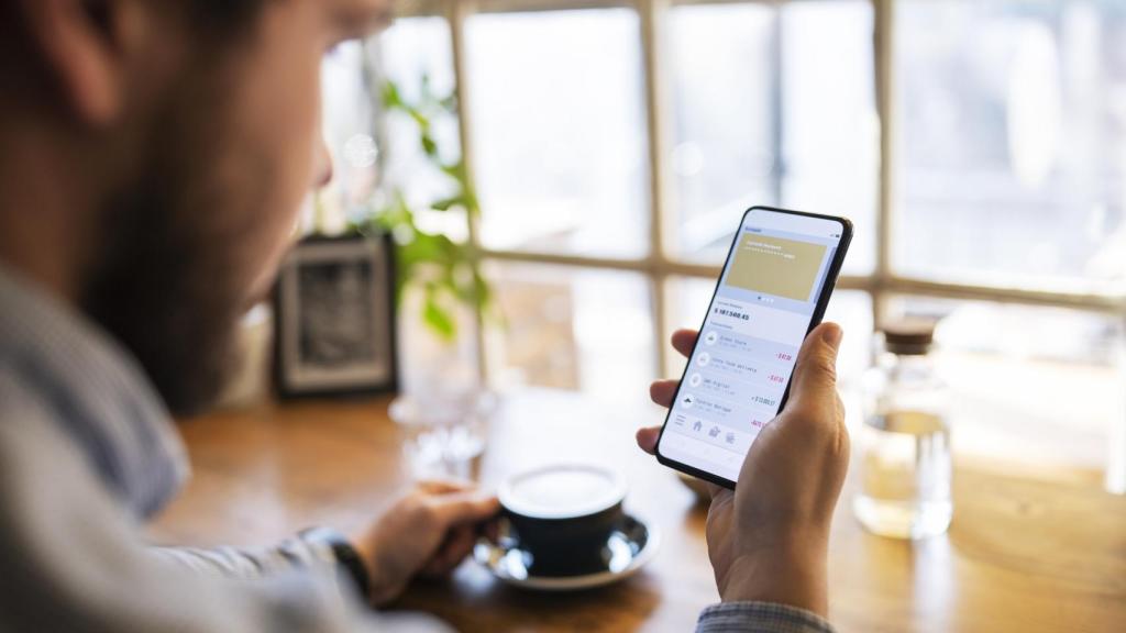 Imagen de un hombre revisa la app de su banco mientras toma un café.