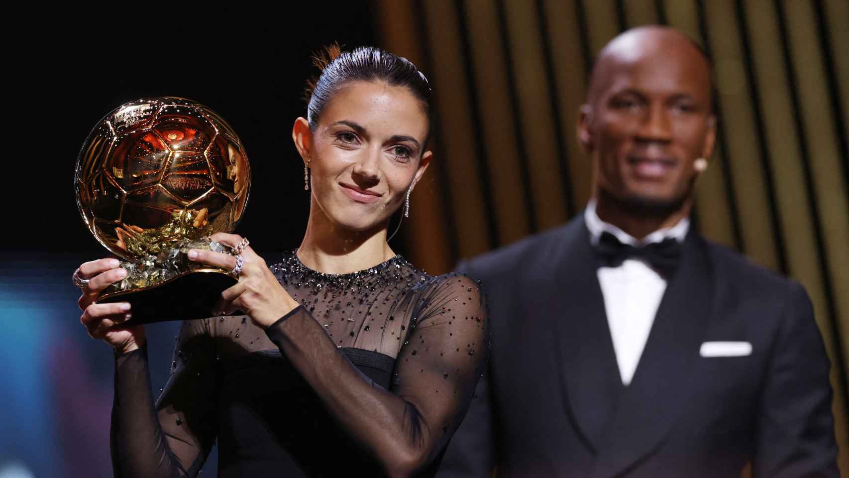 Aitana Bonmatí, con el Balón de Oro y Didier Drogba detrás.