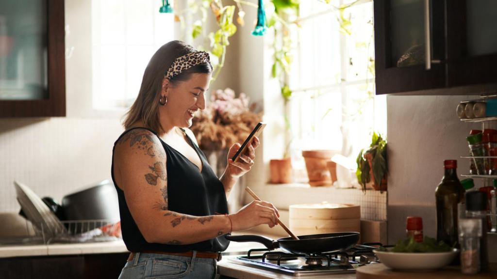 Imagen de una chica joven preparando una tortilla mientras mira el teléfono móvil