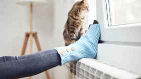 Una persona apoya los pies sobre el radiador mientras su gato mira por la ventana.