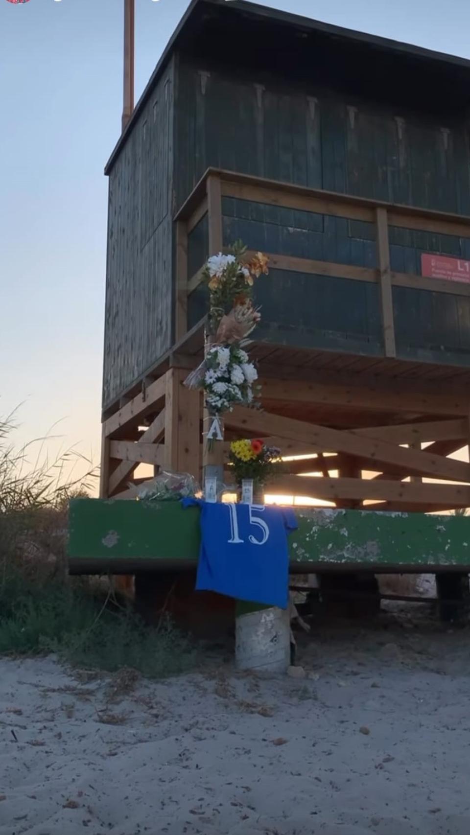 Flores y una camiseta de fútbol sala con el dorsal número 15 que siempre lucía Álvaro bajo los palos, como homenaje de sus amigos en el puesto de socorrismo de El Mojón donde falleció. Justo detrás se observan los pilares habilitados tras la tragedia.