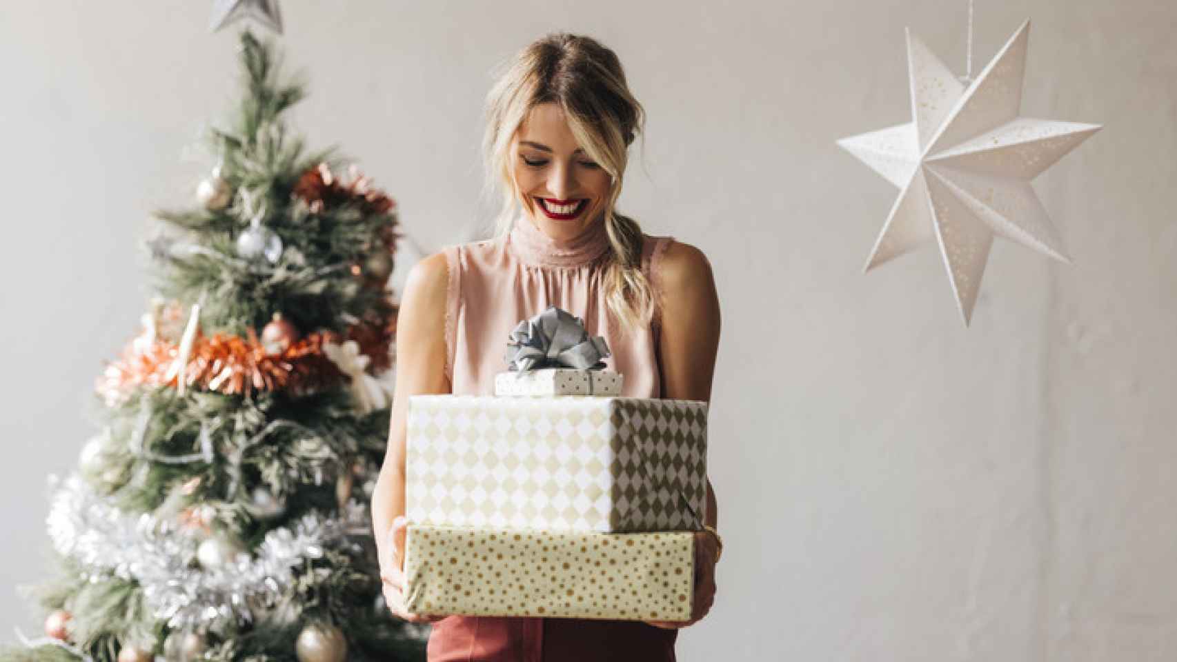 Mujer cargando regalos con un árbol de Navidad en el fondo.