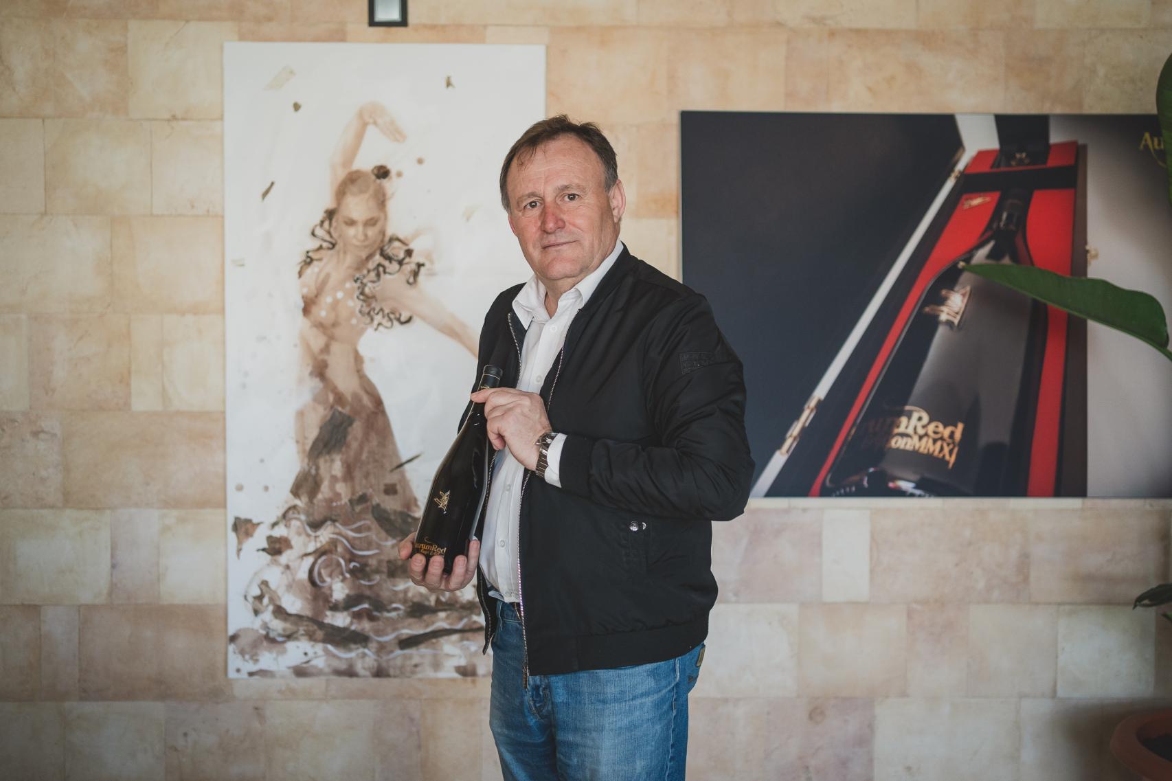 Hilario García, posa en su salón con una botella de AurumRed. Detrás de él, un cuadro pintado sólo con su vino.