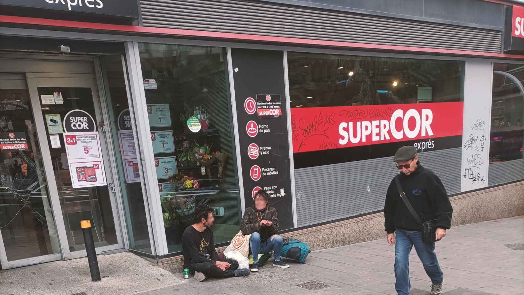 Dos personas sentadas en la calle frente a una conocida cadena de supermercados de Alicante.