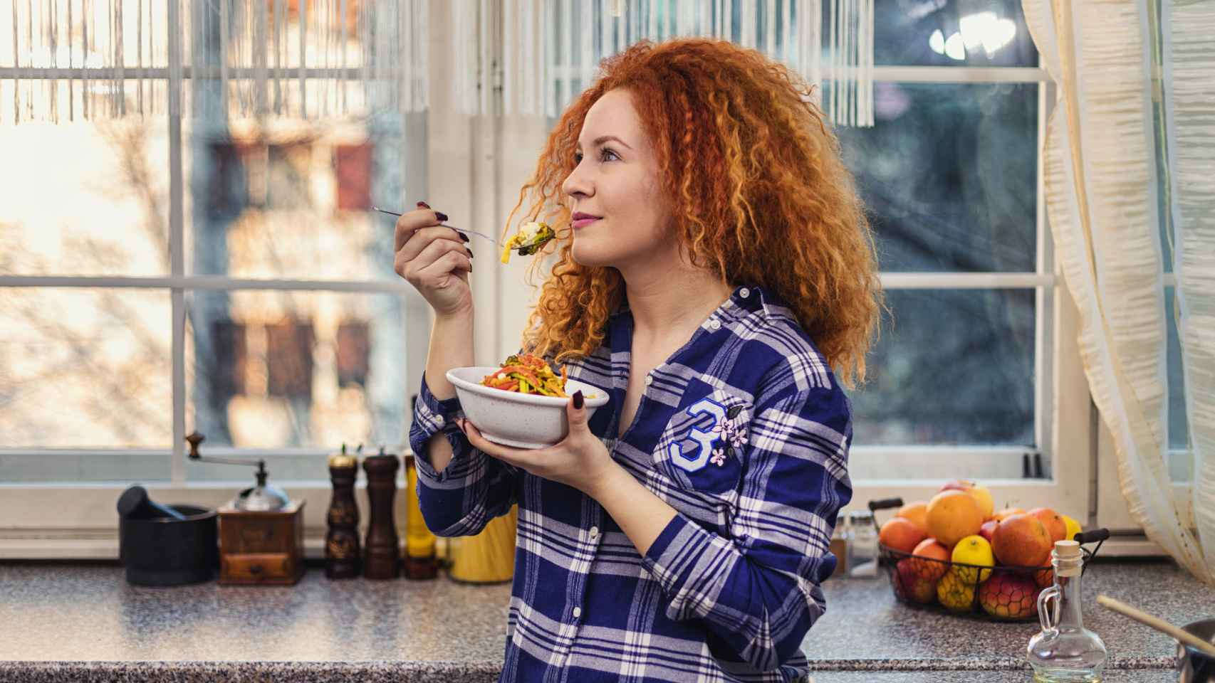 Mujer comiendo de un bol, apoyada en la encima de la cocina.