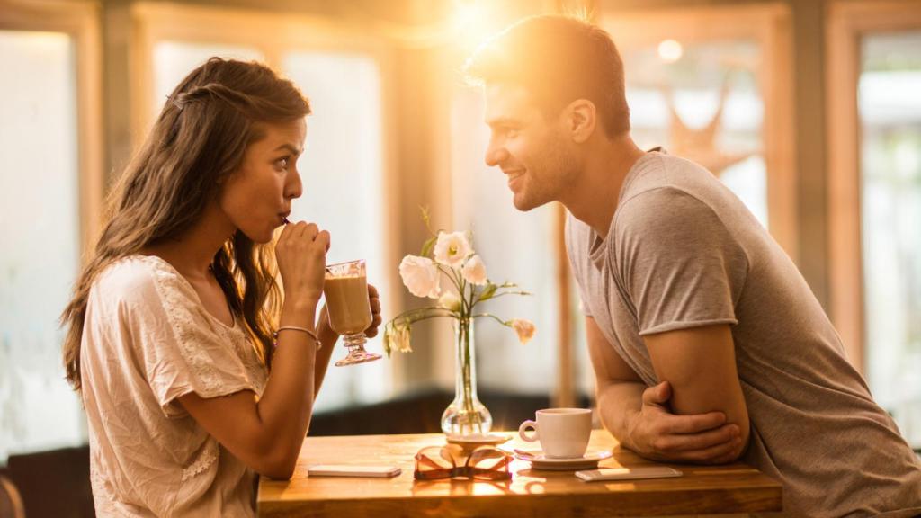 Mujer bebiendo mientras un hombre la mira con atención desde el otro lado de la mesa.