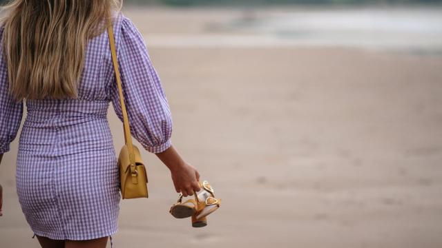 Mujer caminando por la playa con unas sandalias en la mano.