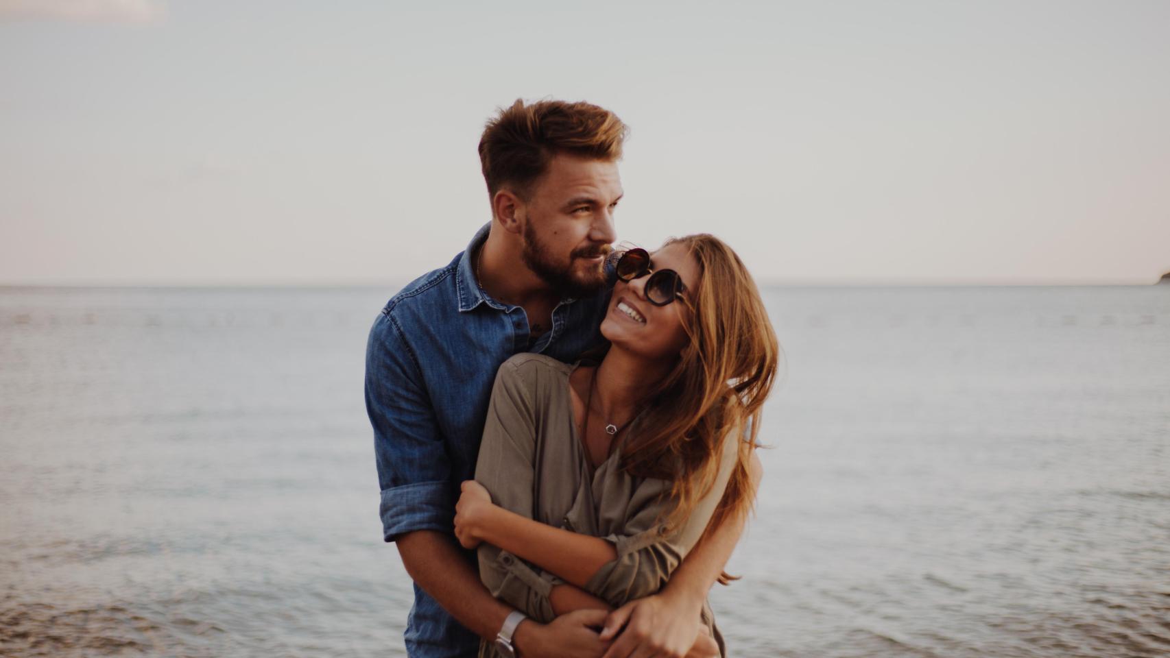 Hombre abrazando a su pareja en la playa con mirada pensativa.