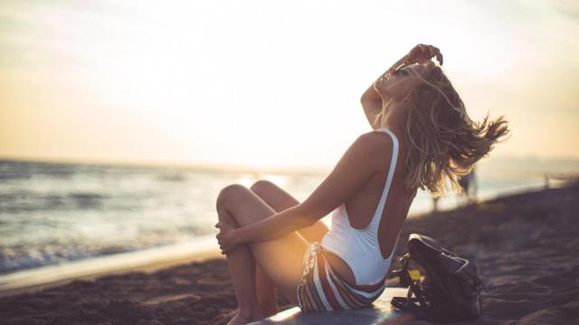 Mujer en la playa sentada sobre una tabla de surf tocándose el pelo.