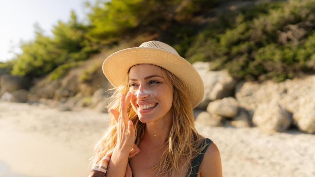 Mujer con sombrero en la playa aplicándose crema solar en la cara.