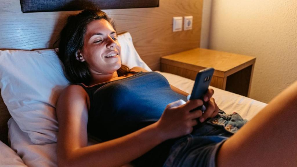 Mujer sonriente sobre la cama mirando el móvil.