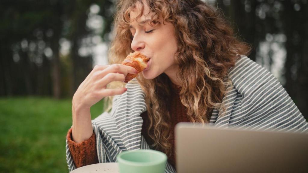 Mujer comiendo un croissant frente al ordenador.