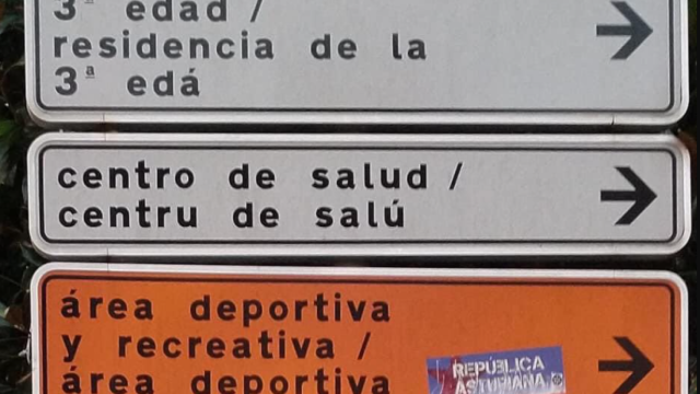 Señales urbanas rotuladas en castellano y en su traducción al asturiano.