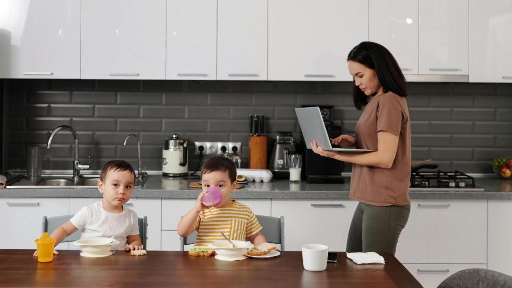 Mujer en la cocina con ordenador en la mano y dos niños sentados a la mesa