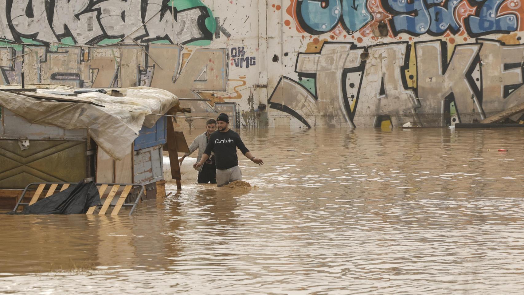 Dos personas vadean un terreno de chabolas inundado junto a la V-30 a causa de las lluvias torrenciales de las últimas horas.