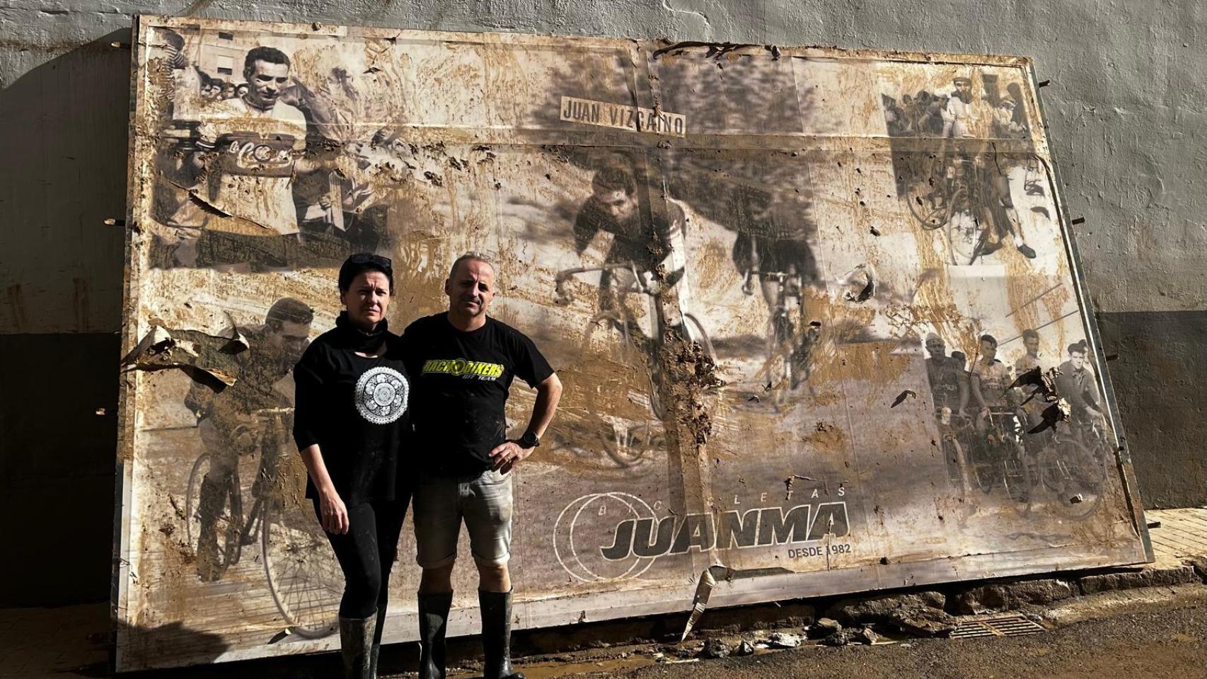 Juanma y su mujer posan frente al cartel de homenaje a su padre, ciclista y fundador del negocio, arrancado del escaparate de su tienda por la DANA.