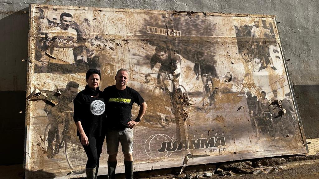 Juanma y su mujer posan frente al cartel de homenaje a su padre, ciclista y fundador del negocio, arrancado del escaparate de su tienda por la DANA.