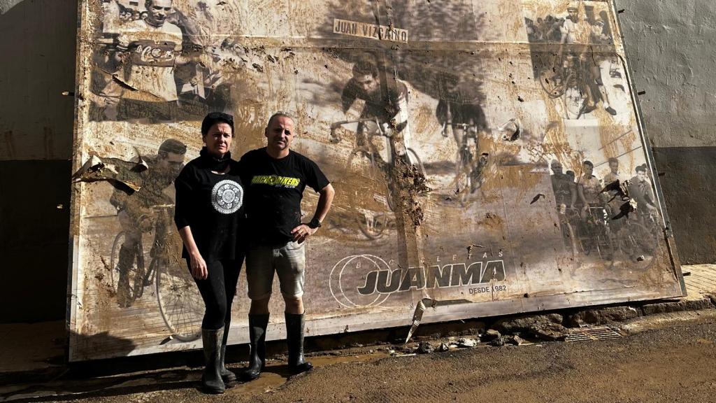 Juanma y su mujer posan frente al cartel de homenaje a su padre, ciclista y fundador del negocio, arrancado del escaparate de su tienda por la DANA.