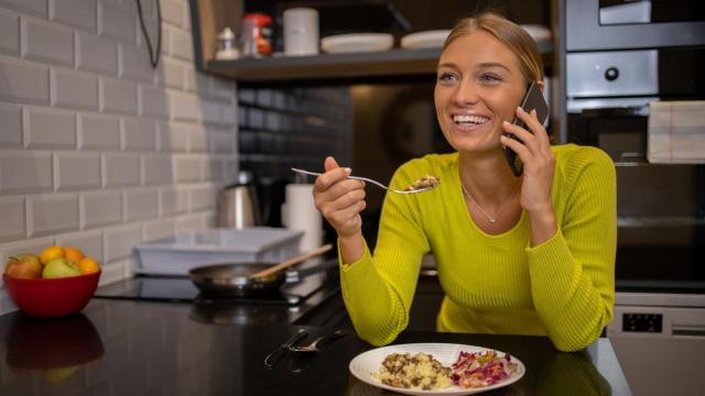 Mujer comiendo en la cocina mientras habla por teléfono.