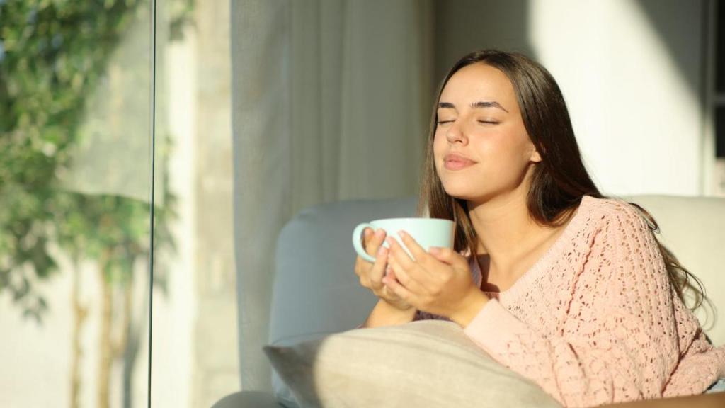 Mujer tomando una taza caliente mientras toma el sol a través de la ventana.